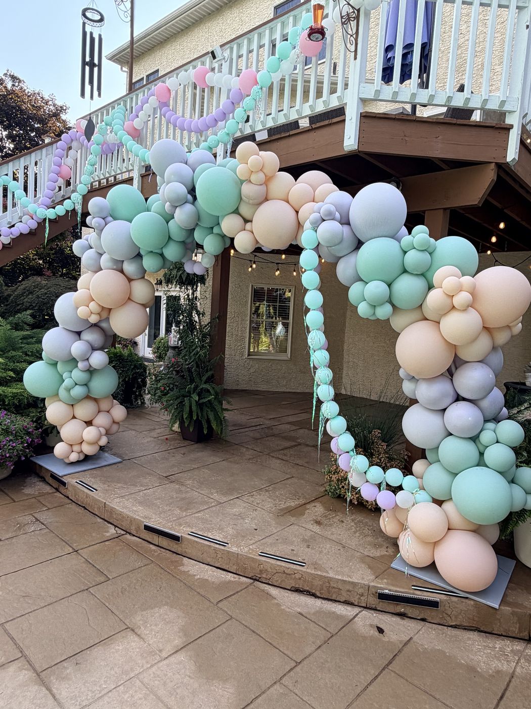 Pastel organic balloon arch in sage, blush, lavender and cream framing a home's lower patio, with a balloon garland along the deck railing